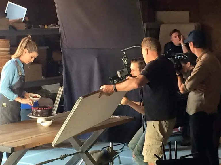 A grip angles a bounce board to shape the light on a tabletop commercial shoot while the camera operator lines up the shot.