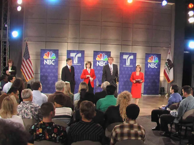 A live televised debate on a lit stage with network banners, flags, a row of candidates, and a seated studio audience.