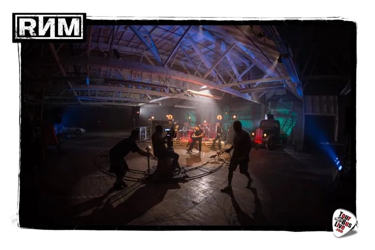 A music-video shoot in a darkened soundstage, a camera operator on a circular dolly track encircling a band under hazy concert lighting.