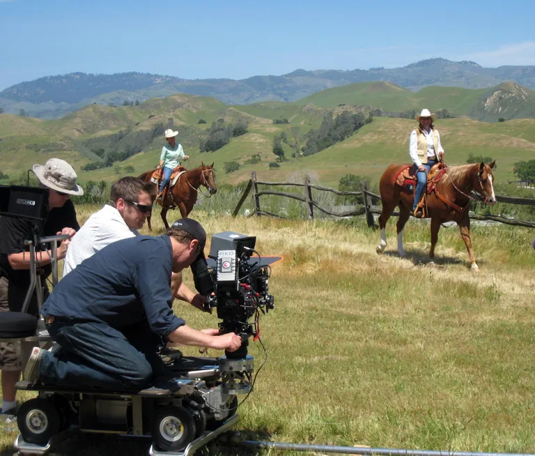 A camera operator on a wheeled dolly and track films two riders on horseback across golden rolling hills.