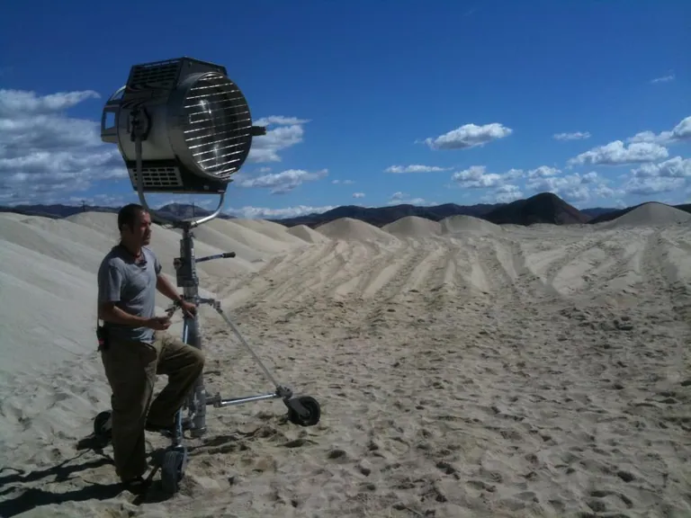 A crew member positions a large 18K HMI fixture on a roller stand in open sand dunes under a clear blue sky.