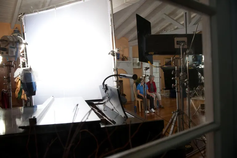 A grand piano interview lit through a large white silk inside a sunlit timber-beam room, cameras and stands in frame.