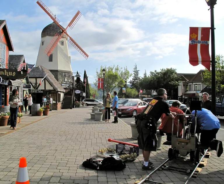 A film crew with a dolly and track shoots on a Solvang street, the village windmill rising behind them.
