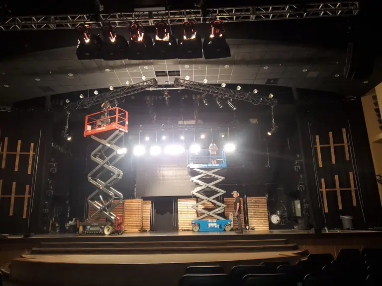 Crew rigging moving-head fixtures on overhead truss from two scissor lifts on a theater stage.