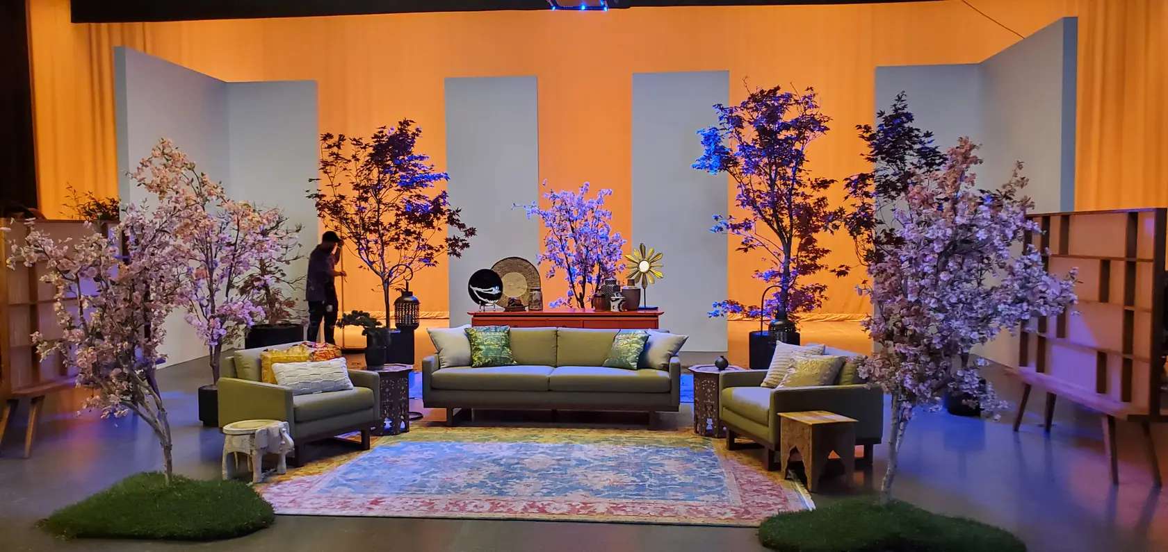 A film set lit with a warm amber wash and cool blue accent light, dressed with faux blossom trees and mid-century furniture.